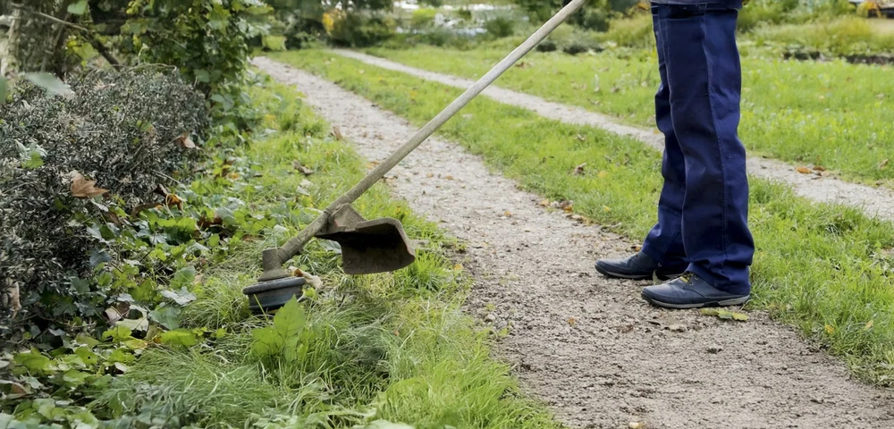 Scène extérieure de maintenance d'espaces verts : ouvrier en combinaison bleue utilisant une houe pour désherber le long d'un sentier gravillonné